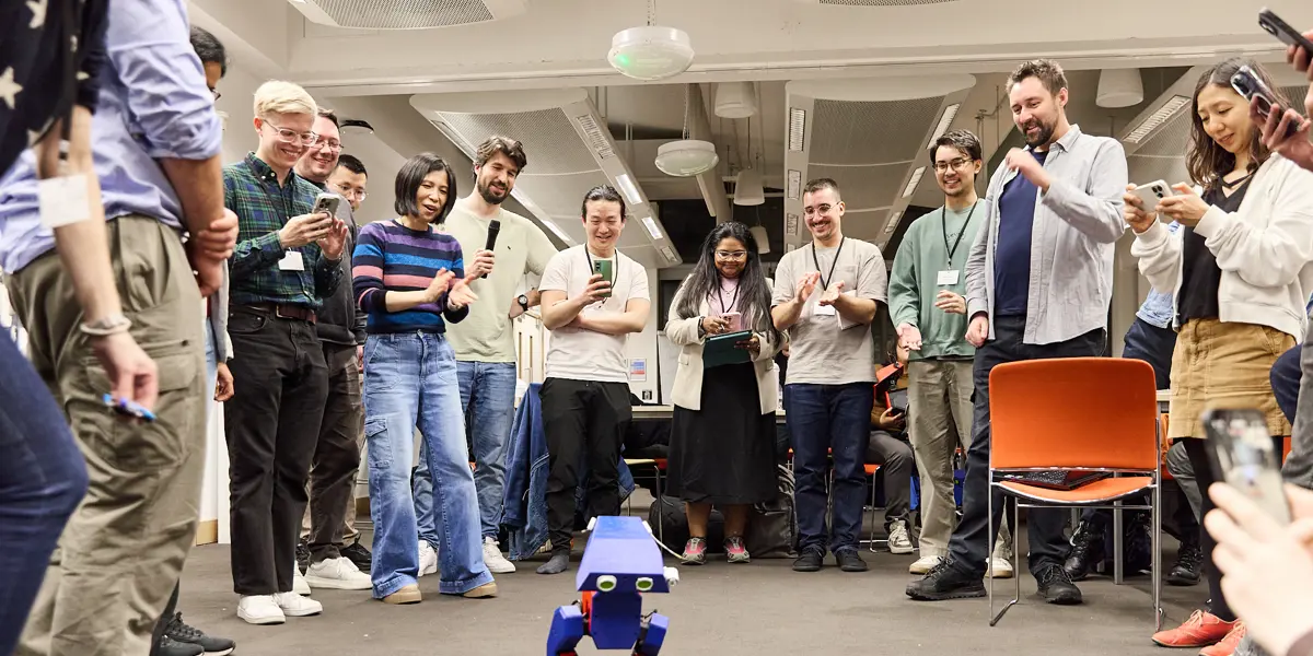 A large group of people standing in a semi circle taking photos and videos of a small blue robot