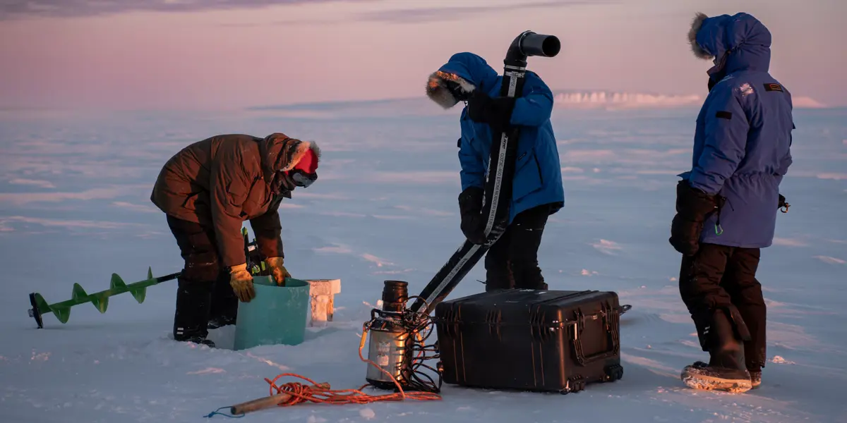 A team of three people dressed in warm winter clothes working on a water pump on ice fields in Canada