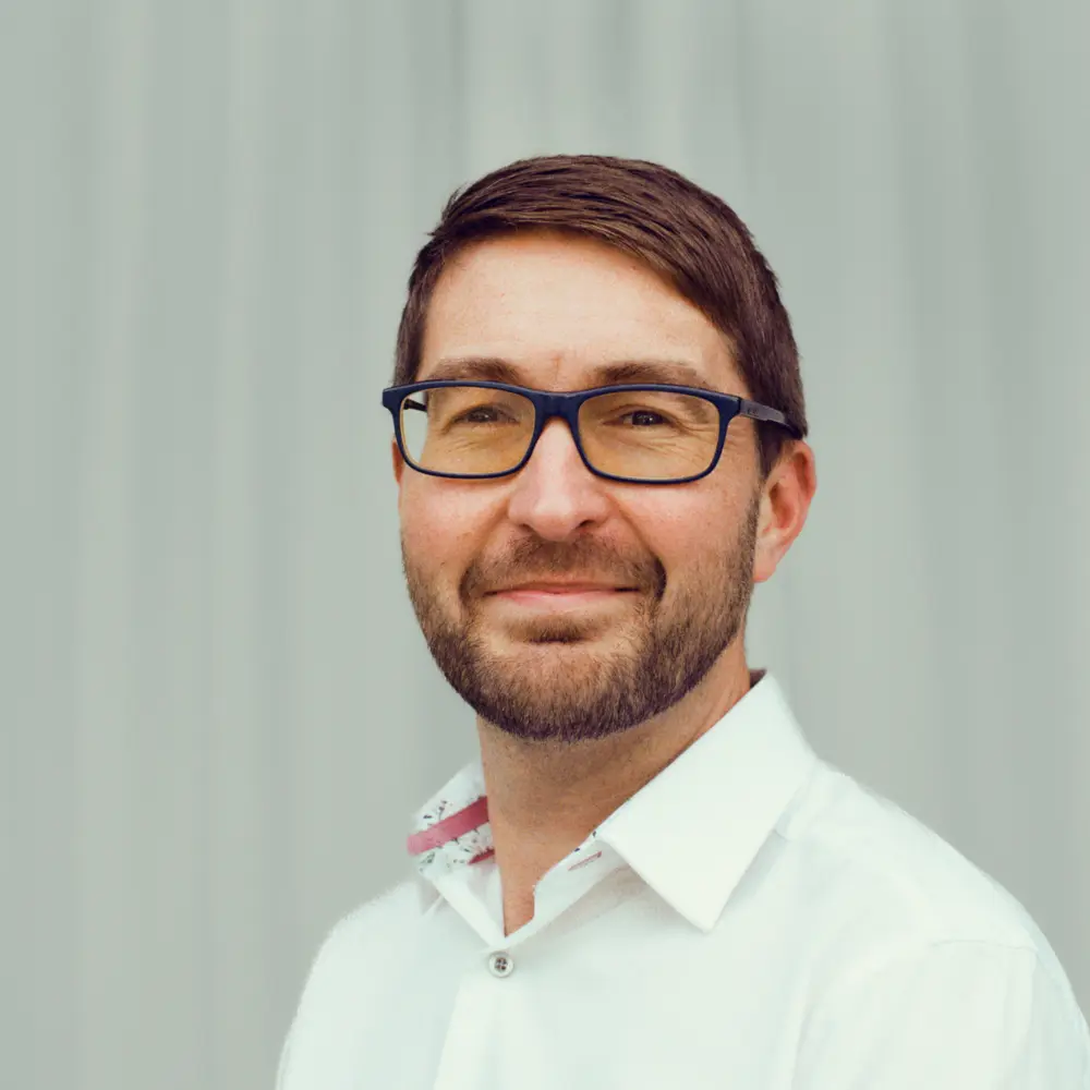 A photo of Mark Symes smiling against a blue wall. 