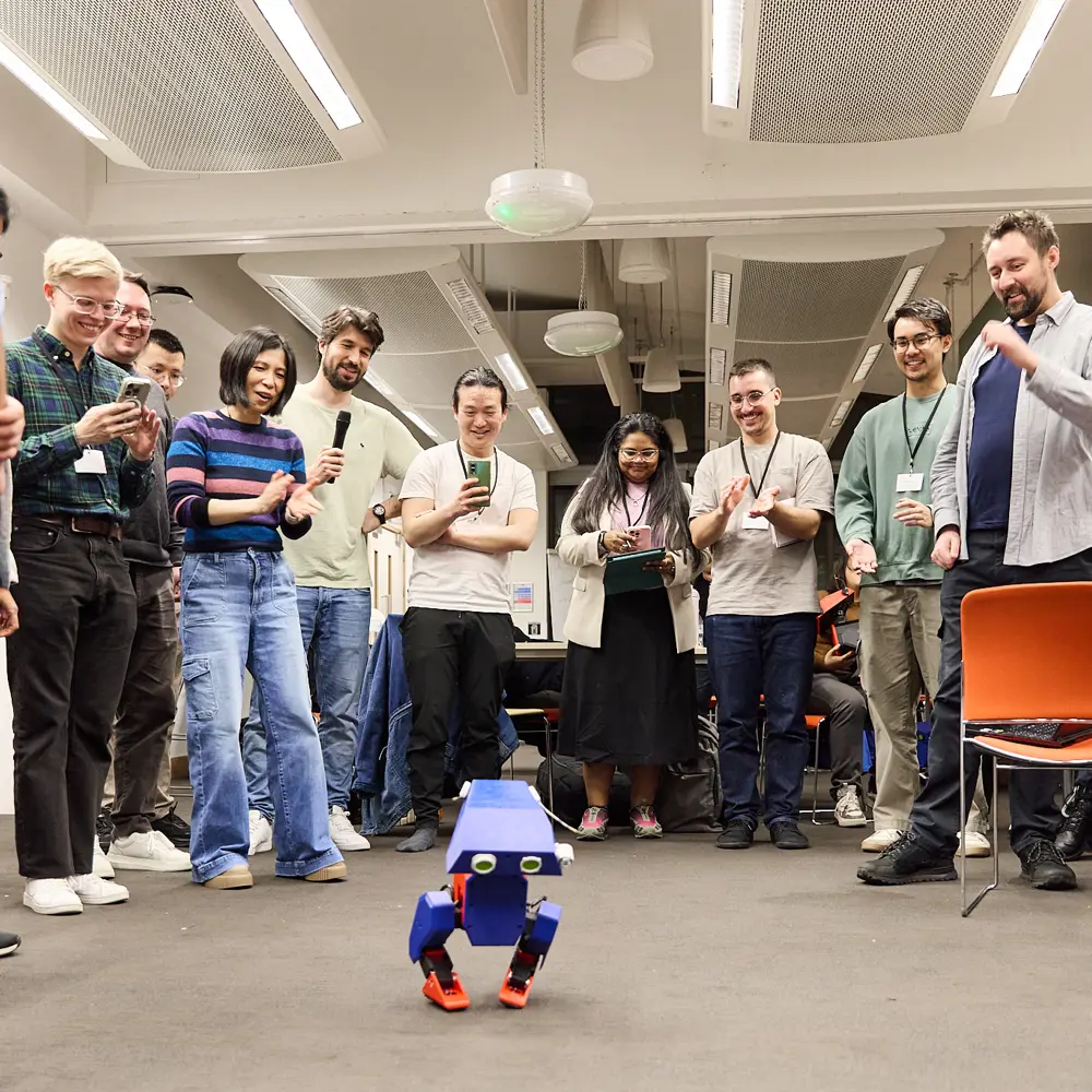 A large group of people standing in a semi circle taking photos and videos of a small blue robot 