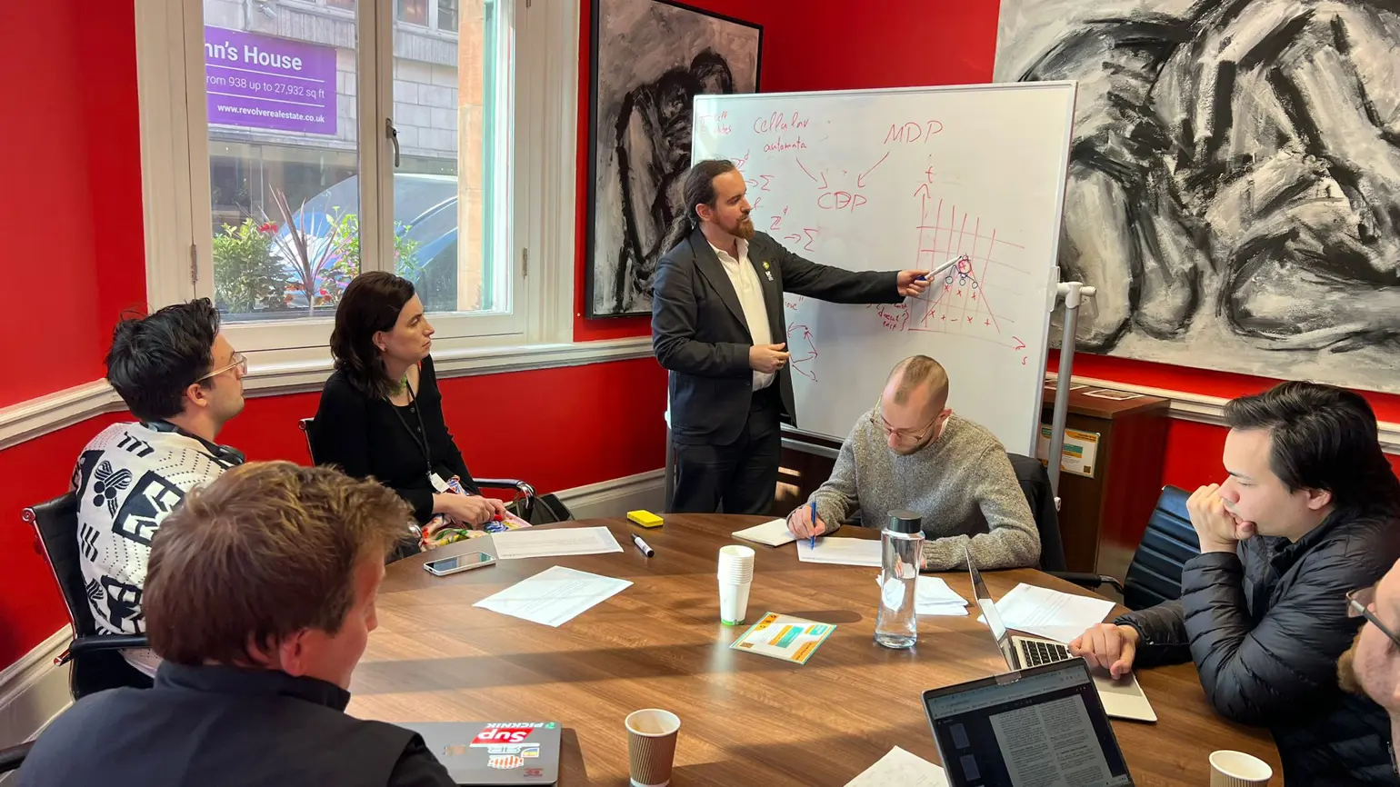 A photo of a group of people sitting around the table and looking at davidad writing on a whiteboard.