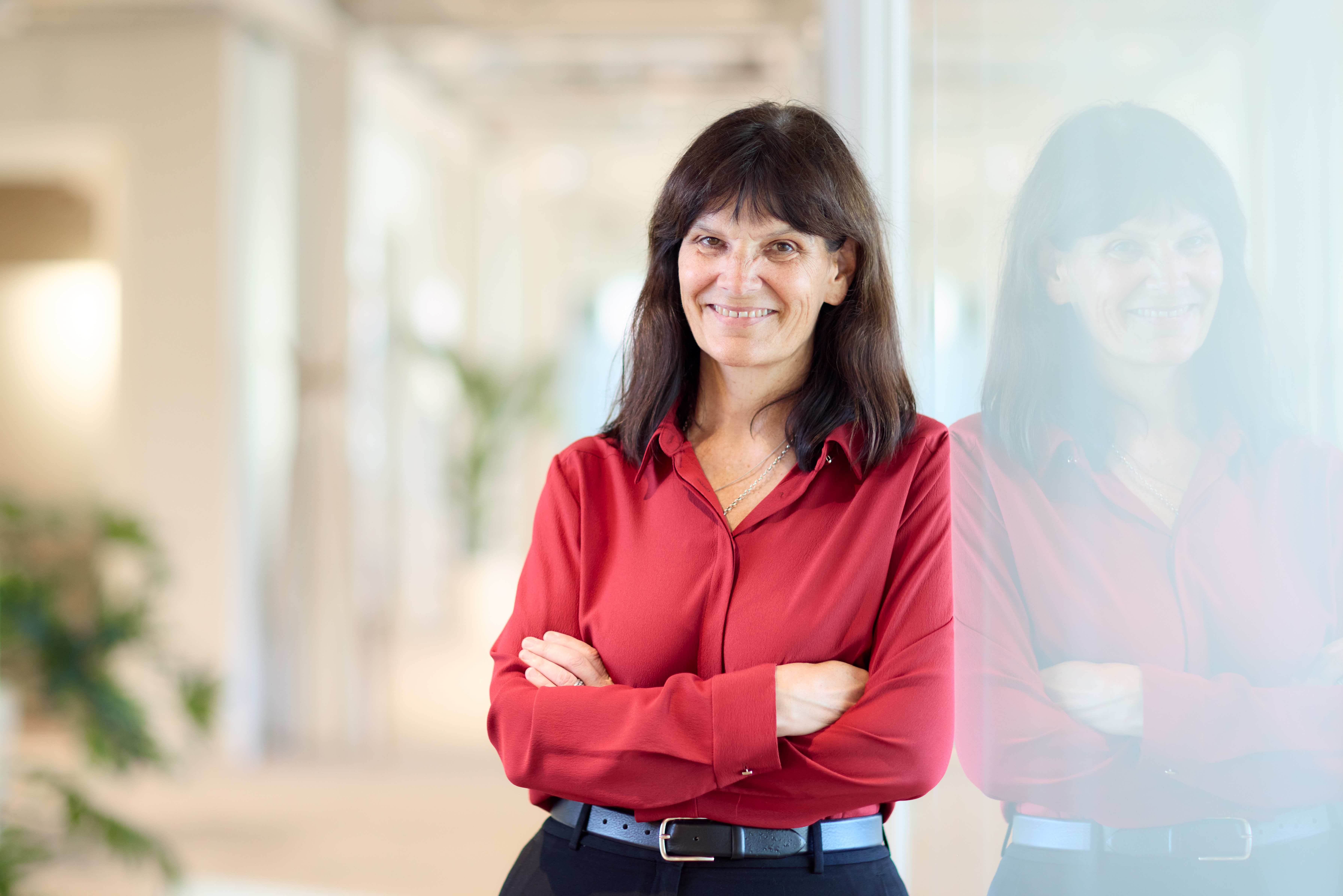 A photo of a woman, Kathleen Fisher, leaning against a glass wall and smiling at the camera with her arms folded. 