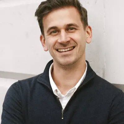 A photo of Fabrizio Ticchiarelli-Marjot smiling at the camera with his arms folded. He is standing against a white wall. 