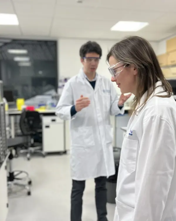 Angie, a Programme Director, in a lab coat and goggles in a lab during discovery. There is also a person in the background speaking and gesturing with their hands. 