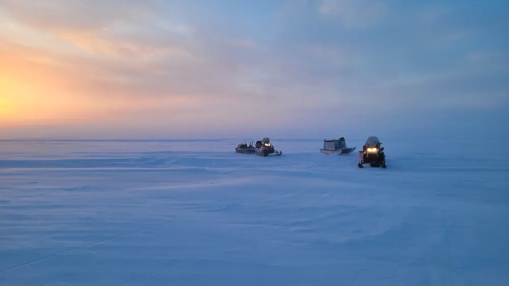 Three snow mobiles sitting on an ice field with a sunset in the background