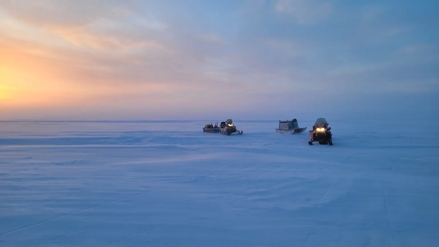 Three snow mobiles sitting on an ice plain with a sunset in the background