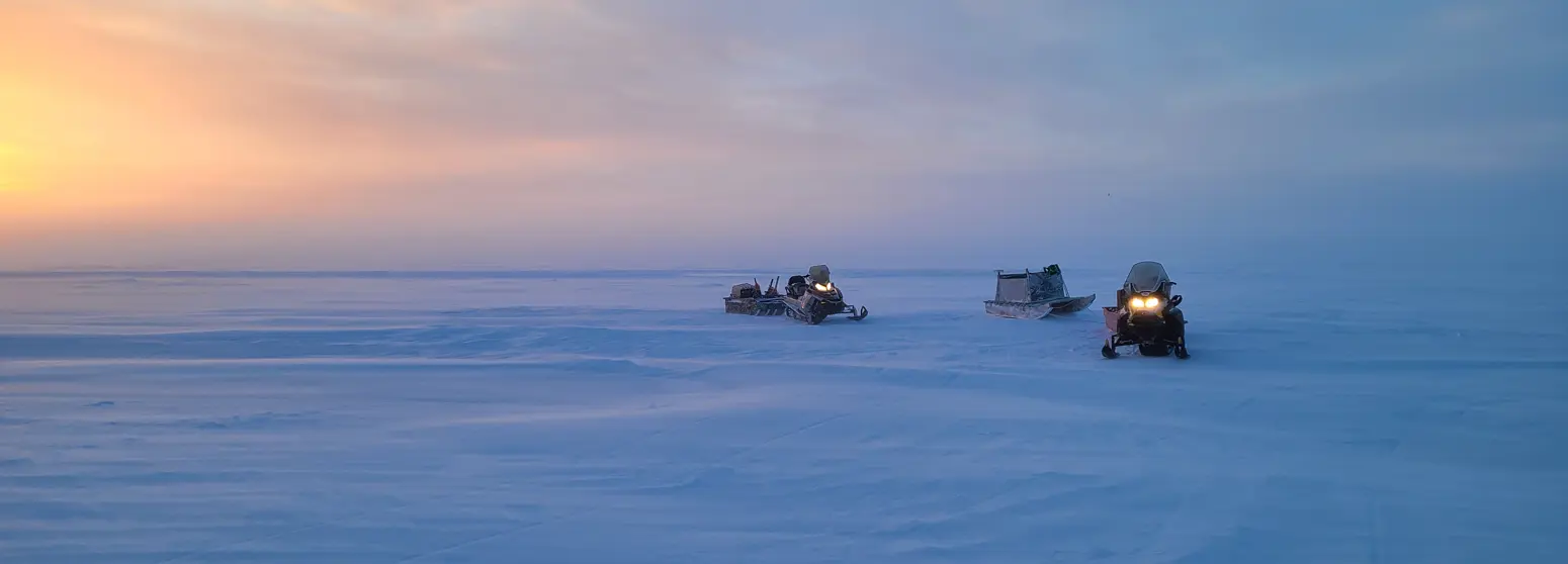 Three snow mobiles sitting on an ice plain with a sunset in the background